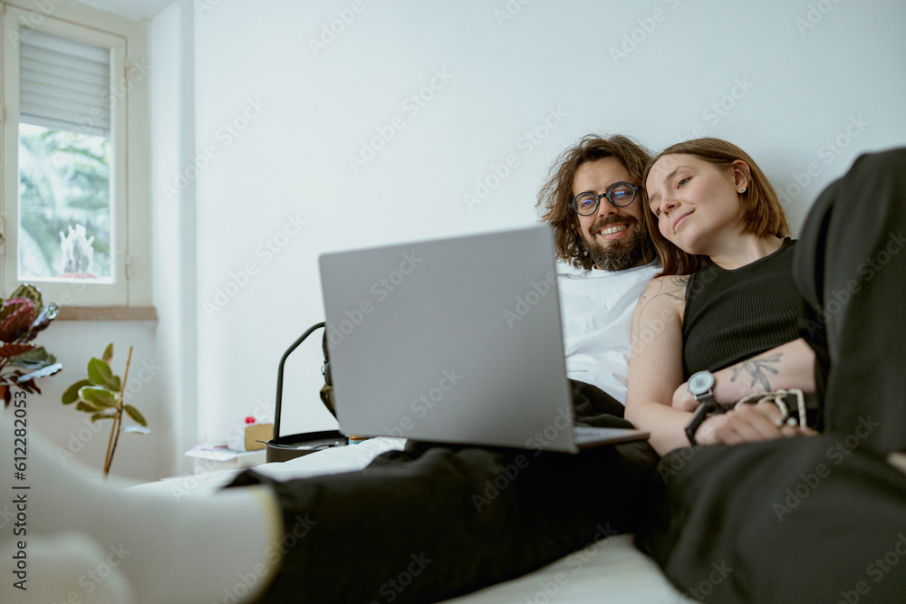 Young family watching TV series using laptop sitting on couch in cozy apartment. Couple in love
