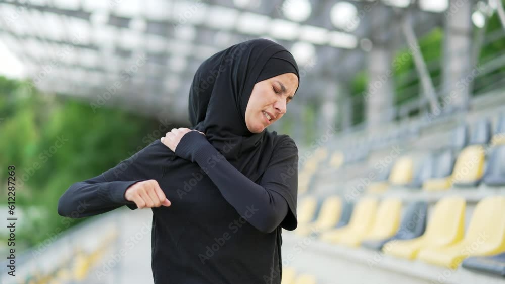 Young sporty muslim woman in hijab feels sharp pain in her shoulder during daytime exercise in a city stadium. Sad female was injured. Unfortunate athlete rubs massages the muscles on the sore spot Young sporty muslim woman in hijab feels sharp pain in her shoulder during daytime exercise in a city stadium. Sad female was injured. Unfortunate athlete rubs massages the muscles on the sore spot