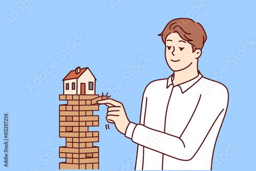 Man puts house in danger by pulling blocks from under building for concept of risky mortgage loan or pledge of real estate. House needs insurance and mortgage redemption from credit bank