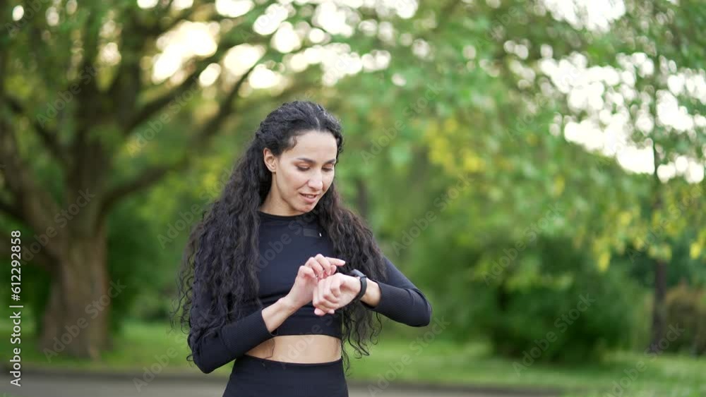 Excited smiling young woman runner looking smart watch in urban city park. Sportswoman using checking fitness tracker. Fit female is satisfied with the results of training, rejoices, shows yes gesture Excited smiling young woman runner looking smart watch in urban city park. Sportswoman using checking fitness tracker. Fit female is satisfied with the results of training, rejoices, shows yes gesture