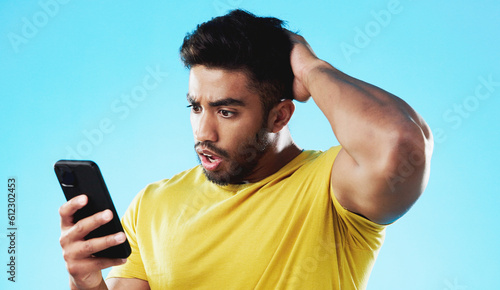 Phone, wow or bad news with a man reading a negative text message in studio on a blue background. Mobile, contact and surprise with a young male looking shocked by a social media post or announcement © Wesley JvR/peopleimages.com