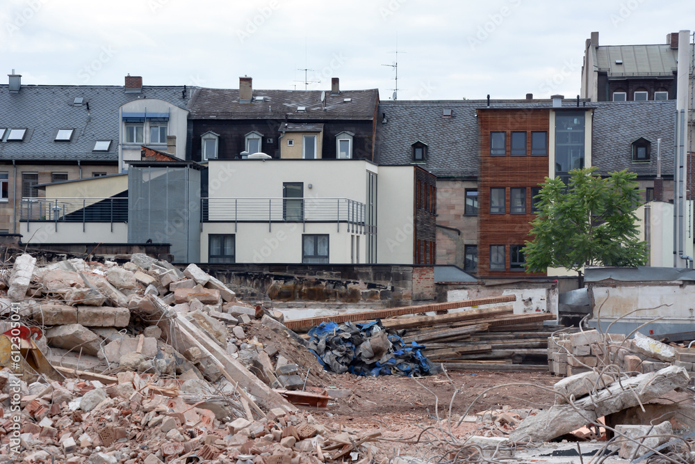 In the foreground are piles of bricks and debris from a destroyed old building. In the background is a row of low-rise buildings