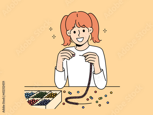 Little girl weaves beaded jewelry sitting at table and creating handmade necklace or bracelet. Elementary school girl with creative hobby creates jewelry and wants to be designer when grows up.
