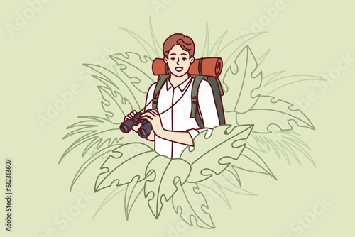 Man traveler stands in thickets of jungle and holds binoculars exploring area in search of further path. Guy traveler with backpack behind back loves adventure and is engaged in hiking