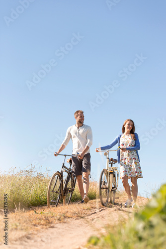 Young couple walking bicycles on dirt road below sunny blue sky