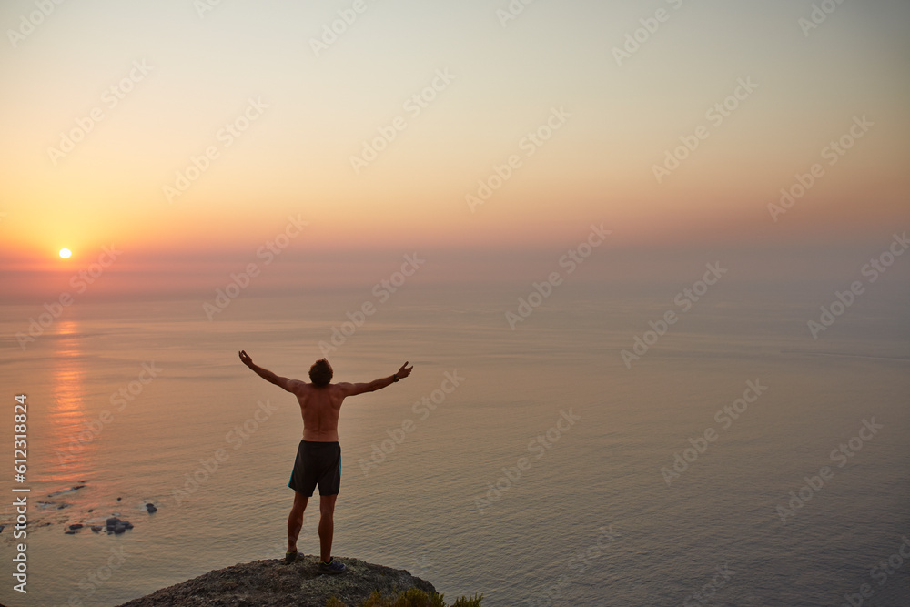 Exuberant male runner arms outstretched on rock overlooking ocean sunset