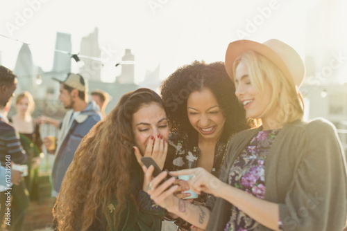 Young women laughing and texting with cell phone at rooftop party