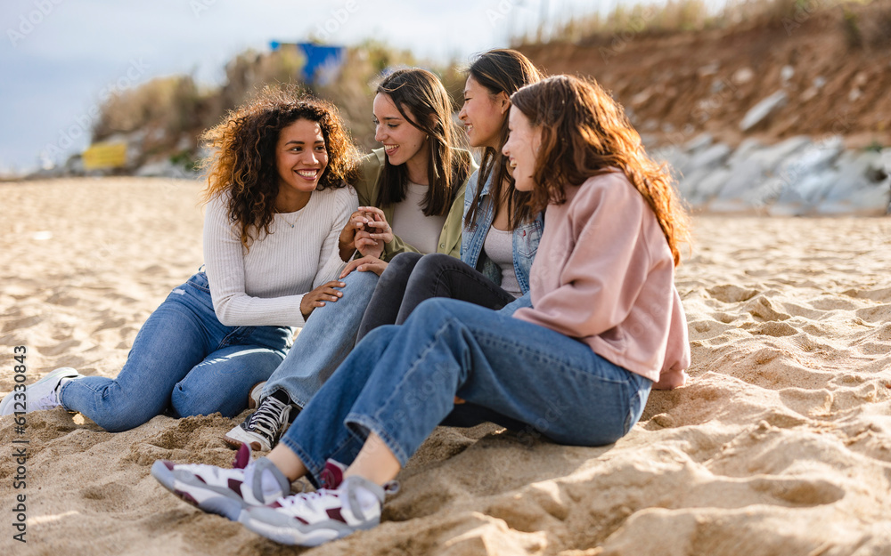 © PintoArt - Young multiracial women laughing out loud on a sunny day - Cheerful group female friends enjoying summer vacation together on the beach sitting down- Summer vacation