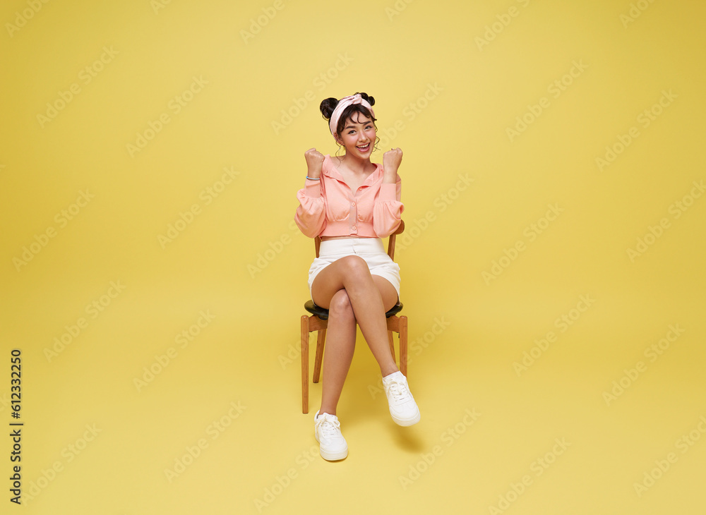 Attractive beautiful Asian woman sitting on chair and hands up raised arms from happiness isolated on yellow background. Excited student woman winner success concept. Attractive beautiful Asian woman sitting on chair and hands up raised arms from happiness isolated on yellow background. Excited student woman winner success concept.
