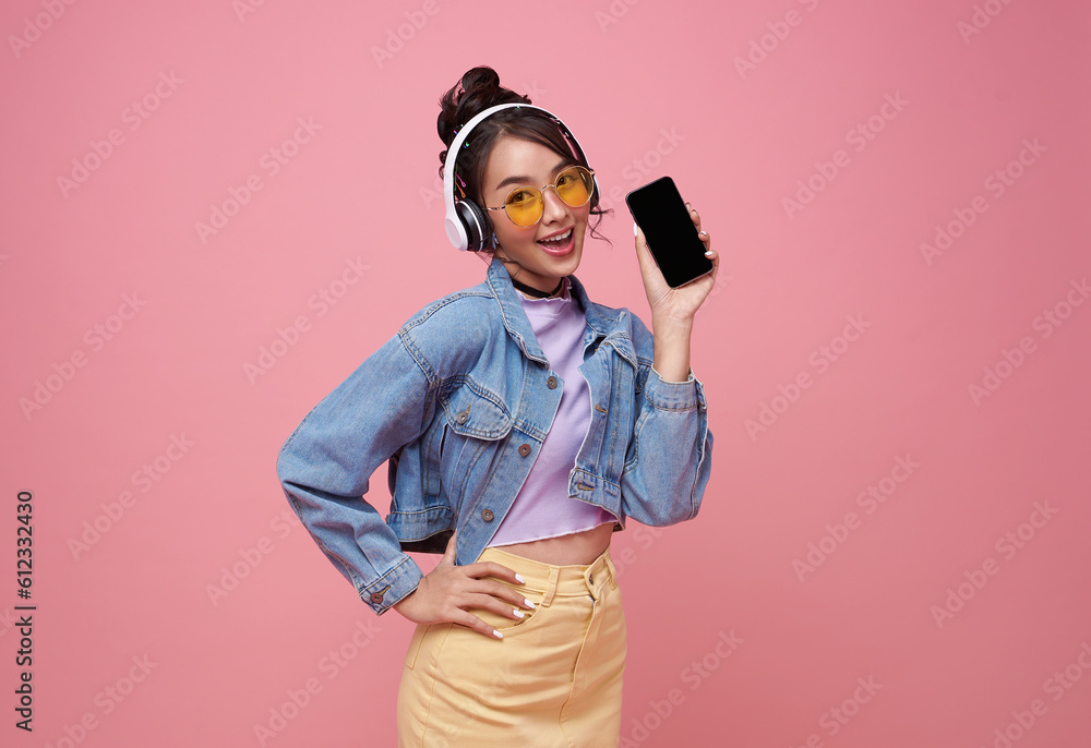 Young Asian teen woman showing smart phone she listening music in headphones isolated on pink background. Young Asian teen woman showing smart phone she listening music in headphones isolated on pink background.
