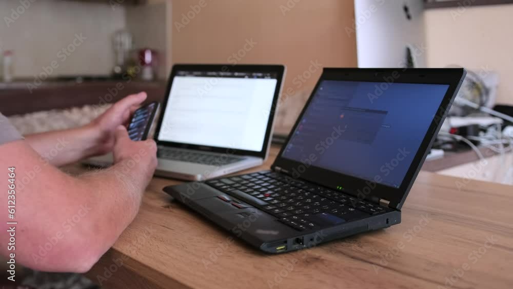 Close-up of male hands that hold smartphone looking through the tape on the screen, man sits at desktop in front of two laptops. Freelancer and work at home concept. Close-up of male hands that hold smartphone looking through the tape on the screen, man sits at desktop in front of two laptops. Freelancer and work at home concept.