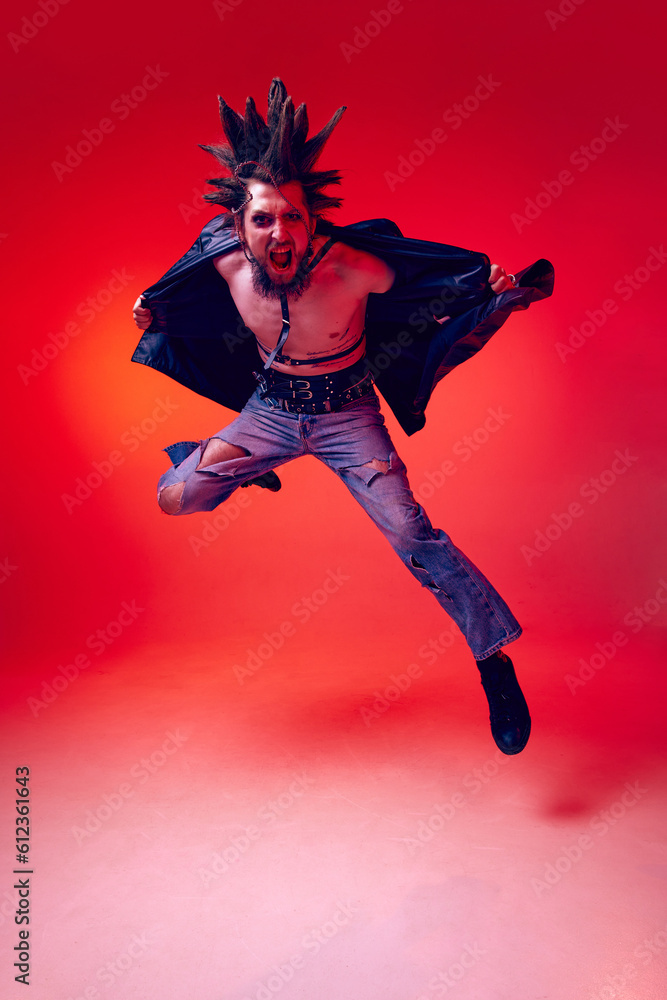 Obraz premium Full-length portrait of young punk in extraordinary clothes, hairstyle and makeup posing over red studio background in neon light. Concept of music, lifestyle, subculture, art, youth, human emotions