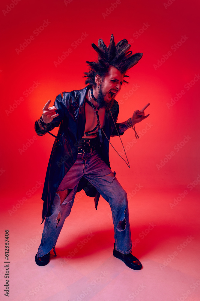 Full-length portrait of young punk in extraordinary clothes, hairstyle and makeup posing over red studio background in neon light. Concept of music, lifestyle, subculture, art, youth, human emotions Full-length portrait of young punk in extraordinary clothes, hairstyle and makeup posing over red studio background in neon light. Concept of music, lifestyle, subculture, art, youth, human emotions
