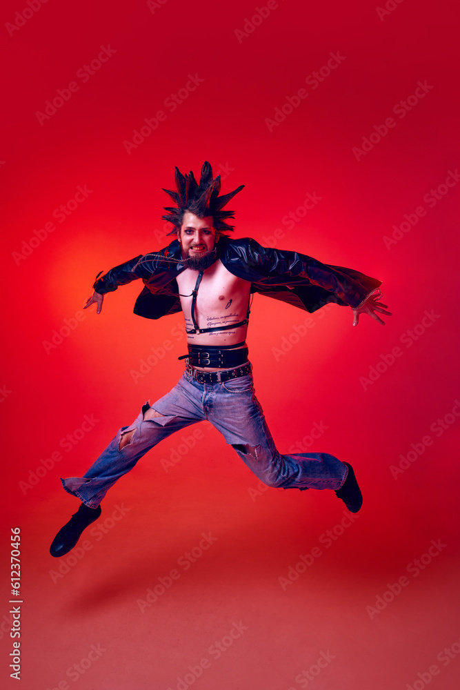 Full-length portrait of young punk in extraordinary clothes, hairstyle and makeup jumping over red studio background in neon light. Concept of music, lifestyle, subculture, art, youth, human emotions
