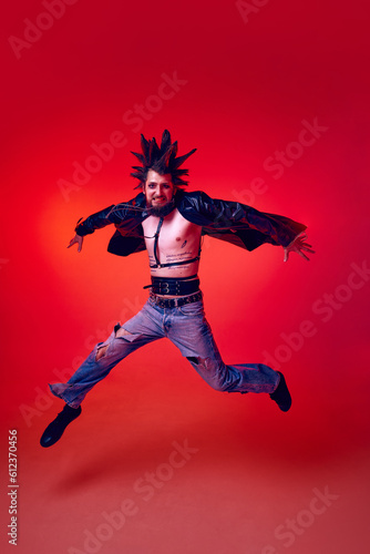 Full-length portrait of young punk in extraordinary clothes, hairstyle and makeup jumping over red studio background in neon light. Concept of music, lifestyle, subculture, art, youth, human emotions