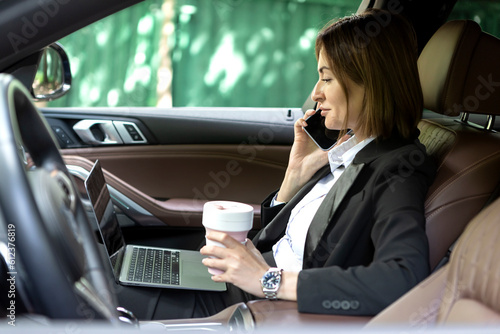 Successful attractive business woman sitting in the passenger seat in her luxury car working on laptop, holding a hot coffee mug and talking to mobile phone about business meeting © Vitaliy