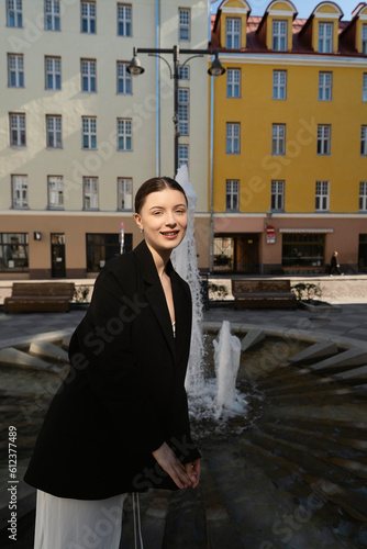 Beautiful young brunette in white dress and black blazer walking outdoors on the street of an European city against a fountain and fancy old buildings