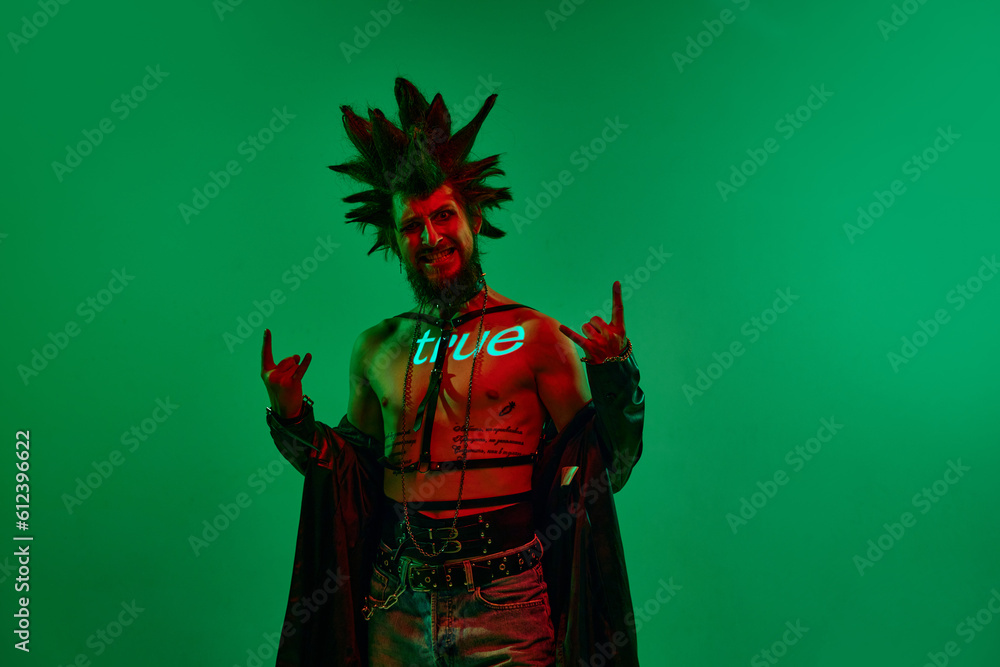 Fototapeta premium Expressive young man, rock musician, punk with neon filter on body posing against green studio background in neon light. Concept of music, lifestyle, subculture, art, youth, human emotions