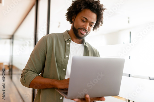 Smart and intelligent African-American businessman wearing smart casual green shirt using laptop indoors, male office employee holding computer and typing message, smiling, enjoying his job