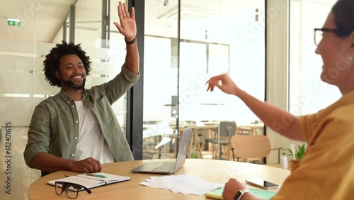 Two young happy diverse colleague sitting at desk and taking high five while working together in coworking space, man and woman celebrating successful project completion or good deal