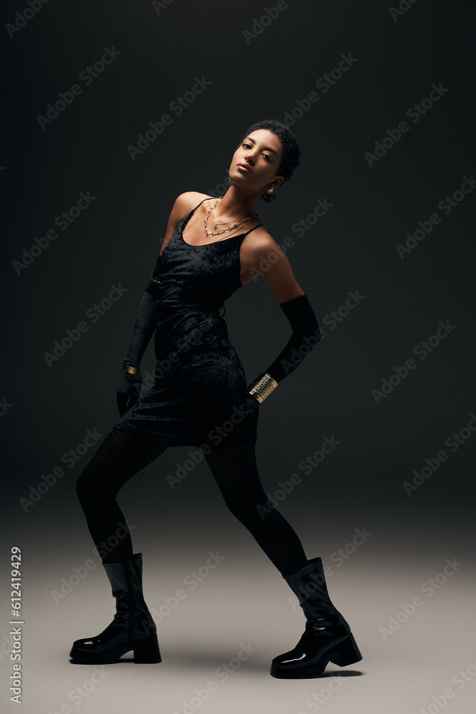 Full length of trendy african american woman in little dress, gloves and stylish boots posing while looking at camera and standing on black background, high fashion and evening look