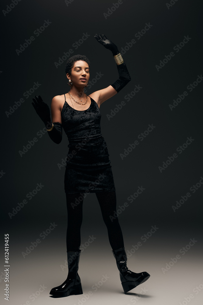 Full length of elegant african american model in little dress, gloves and golden accessories standing with closed eyes on black background, high fashion and evening look Full length of elegant african american model in little dress, gloves and golden accessories standing with closed eyes on black background, high fashion and evening look