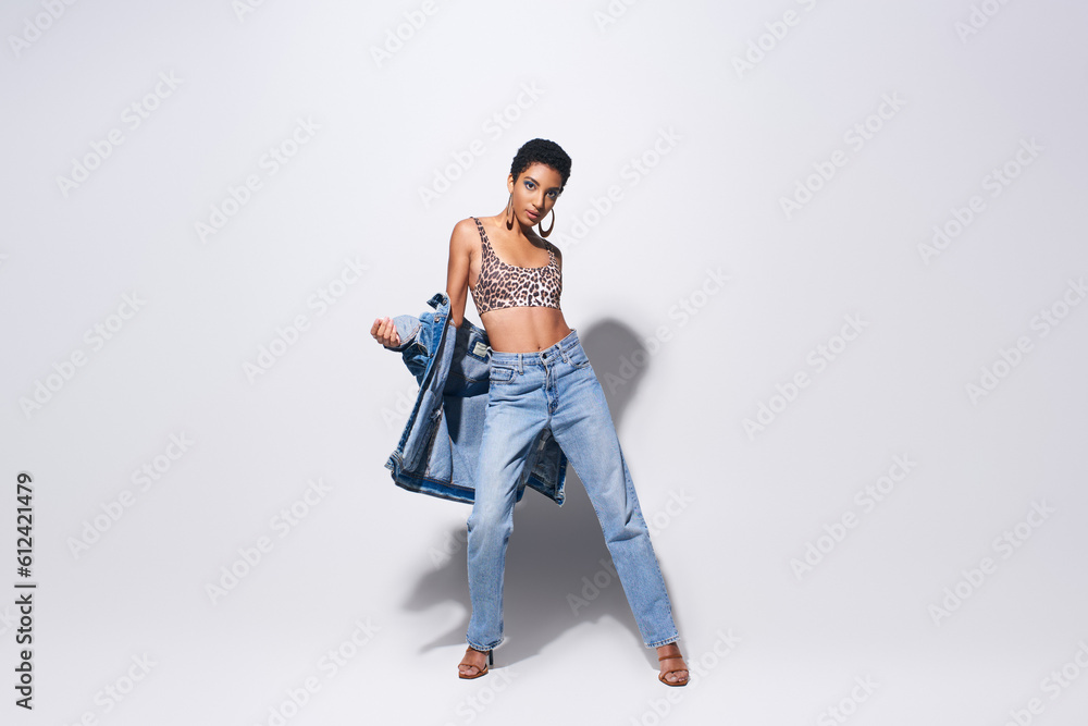 Naklejka premium Full length of fashionable african american woman in top with animal print, denim clothes and heels posing and standing on grey background, denim fashion concept