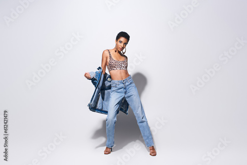 Full length of fashionable african american woman in top with animal print, denim clothes and heels posing and standing on grey background, denim fashion concept