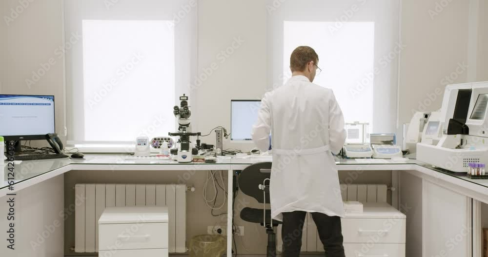 Male doctor analyzing samples in lab. Handheld shot of man in white robe with gloves and glasses putting vial with sample into machine during work in medical laboratory in clinic. Male doctor analyzing samples in lab. Handheld shot of man in white robe with gloves and glasses putting vial with sample into machine during work in medical laboratory in clinic.