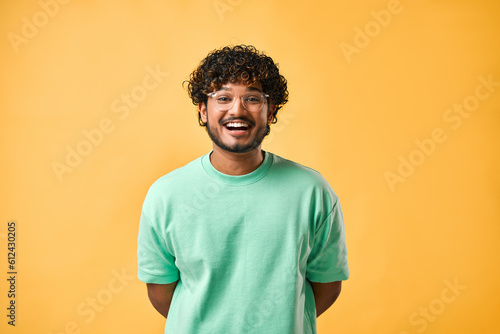 Candid portrait of a handsome curly-haired young man in a turquoise t-shirt and glasses on a yellow background laughing.