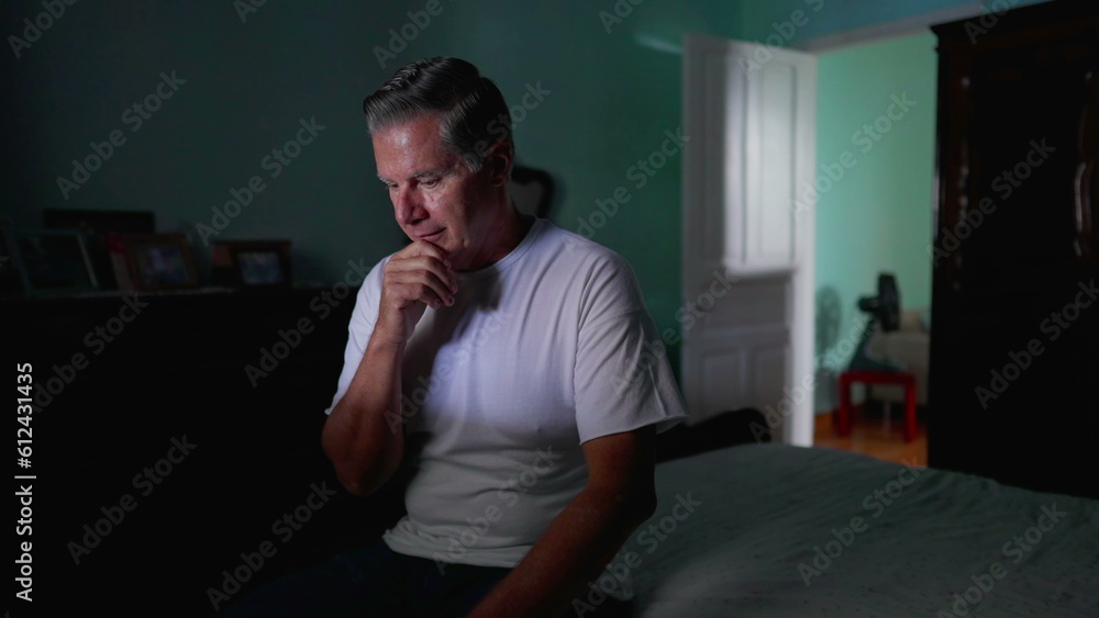 Fototapeta premium Thoughtful older man sitting by bedside with pensive expression. Domestic lifestyle of middle-aged person pondering solution to problem