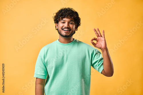 Handsome indian man in turquoise t-shirt showing ok gesture with one hand while looking at camera and smiling while standing against yellow background. photo