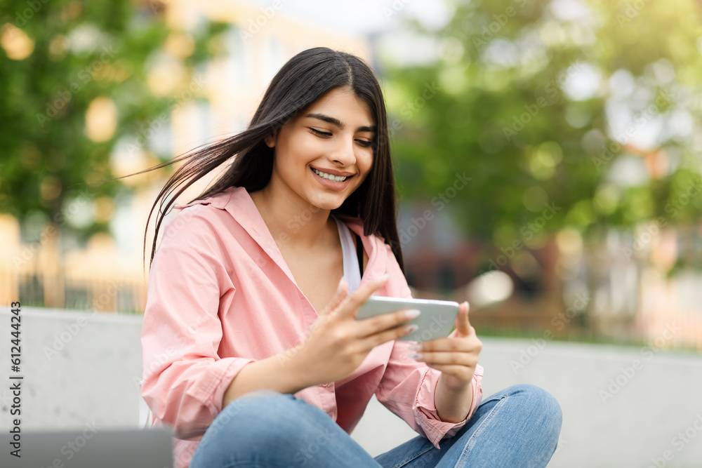 Smiling teen hispanic lady messaging or playing on smartphone while relaxing in park outdoors, free space Smiling teen hispanic lady messaging or playing on smartphone while relaxing in park outdoors, free space