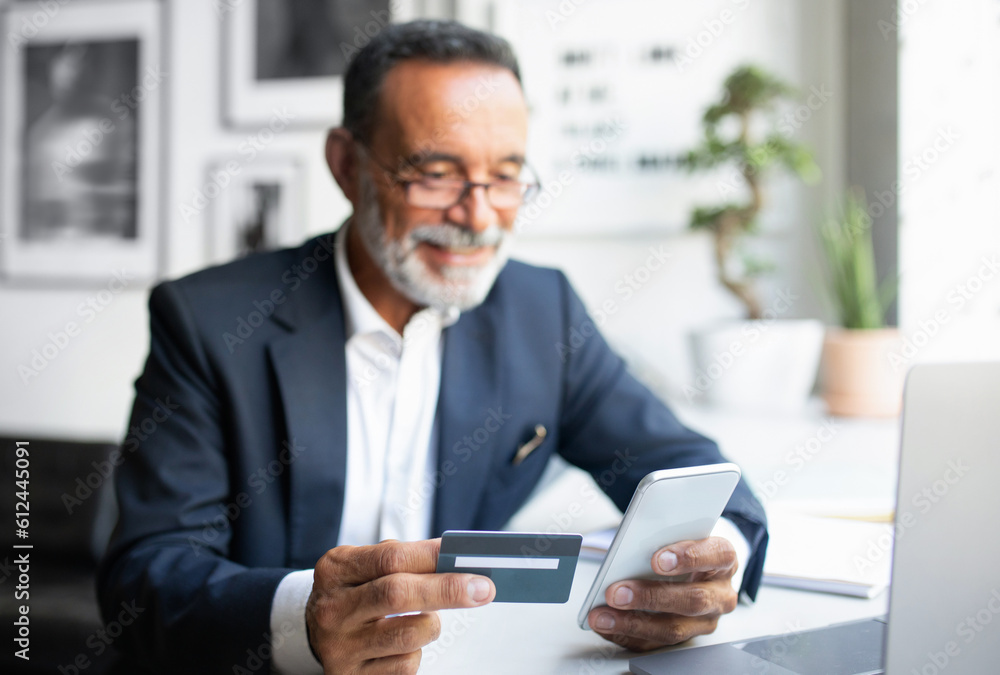 Cheerful european senior businessman in suit, glasses with laptop use phone, credit card at table in office Cheerful european senior businessman in suit, glasses with laptop use phone, credit card at table in office