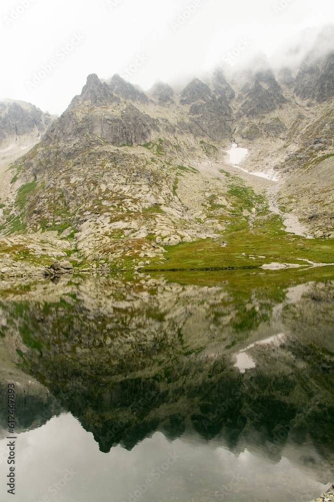 Scenic view of a stone mountains covered by snow and green grass and a reflection of it on a lake