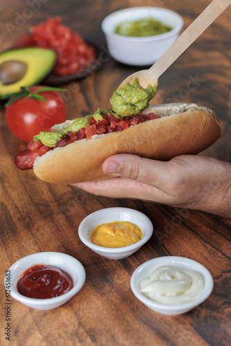 Completo italiano: vertical. Adding palta, with wooden spoon, on tomato and vianesa on hot dog bun, one hand, tomato, avocado, mayonnaise and mustard on a wooden table. Typical Chilean food concept