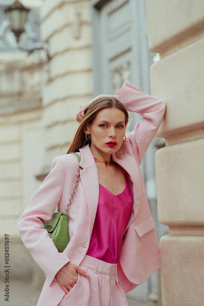 Fototapeta premium Fashionable elegant confident woman wearing trendy pink suit blazer, satin top, heart necklace, trousers, with green faux leather shoulder bag, posing in street. Outdoor fashion portrait