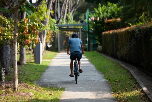 Young singaporean Man with blue t-shirt riding a bike in a narrow road surrounded by greenery © Mathieu Gallon/Wirestock Creators