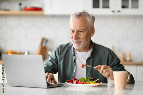 Smiling Senior Man Using Laptop And Having Breakfast In Kitchen