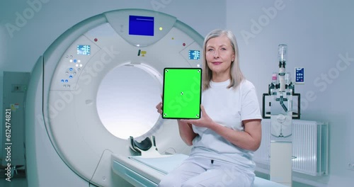 Mature woman is sitting at TC scanner bed. Female at room of MRI. Woman dressed up in white is showing screen of chroma key tablet. Female patient is posing at background of medical equipment. photo