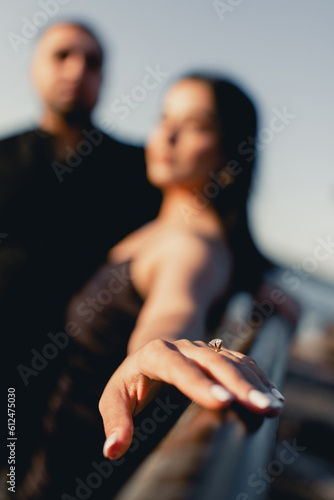 An engagement ring with a precious stone on the finger of a woman who is hugged by her man. A young couple in love is dressed in black and posing at sunset. The man made a marriage proposal