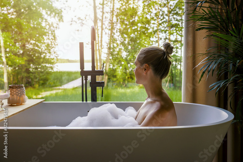 young woman relaxing in bathtub full of foam and looking out of window at home bathroom