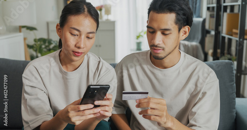 Happy young Asian couple sit on couch spend time together have fun use smartphone devices shopping use credit card online payment at home. Young husband and lifestyle domestic activity concept.