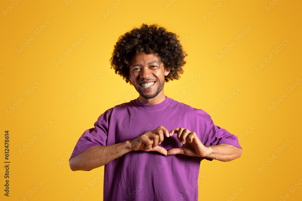 Cheerful black adult curly man in purple t-shirt makes heart gesture with hands
