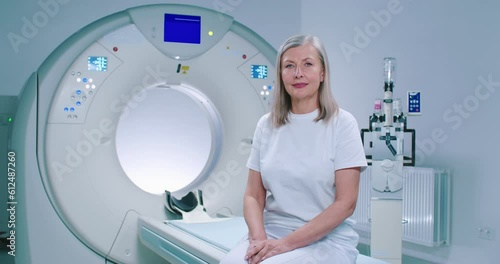 Shooting with smooth zoom. Mature woman sits at TC scanner bed. Female at room of MRI. Woman dressed up in white is looking at camera. Female patient is posing at background of medical equipment. photo