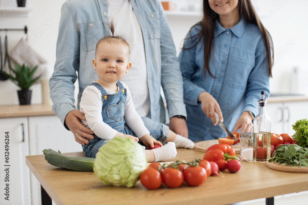 Fototapeta premium Close up view of adorable infant sitting on dining table among fresh crunchy products ready for garden salad. Cute baby daughter participating in family dinner preparation on sunny day at home.