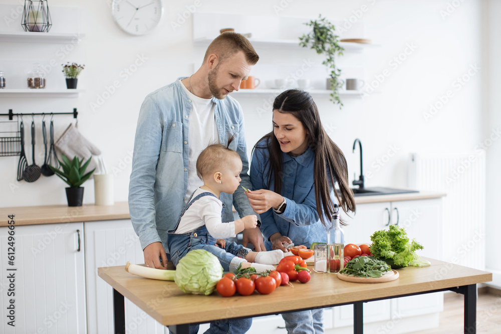 Caring young woman in denim clothes letting baby girl taste green vegetable while cooking dishes for dinner at noon. Mindful parents getting child used to new textures and new flavours of solid food.