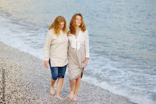 Redhead senior mother and her adult beautiful pregnant daughter are walking together on the sea shore. © Maria Sbytova
