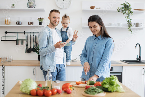 Photography Smiling blond man using cell phone while standing near countertop with infant girl in home kitchen