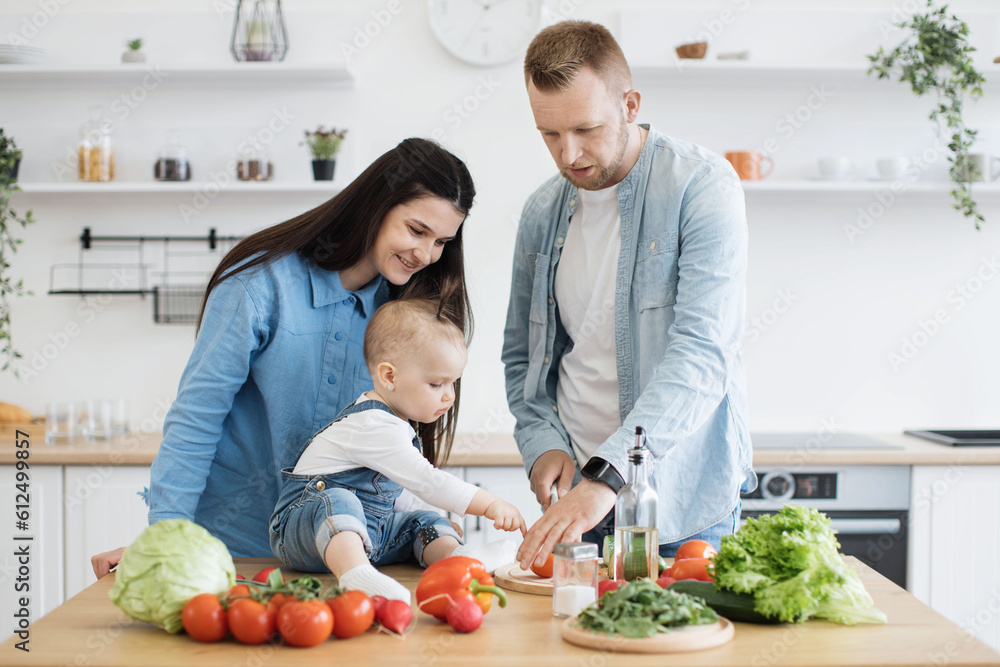Curious small child in cozy wear pointing at tomato on cutting board while attentive man slicing vegetable for salad. Caring mom and dad encouraging baby girl to understanding safety rules on kitchen.